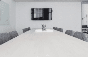Ein Büro mit einem großen hellgrauen Holztisch um den 5 graue Stühle stehen. An der gegenüberliegenden Wand hängt ein ausgeschalteter Fernseher.